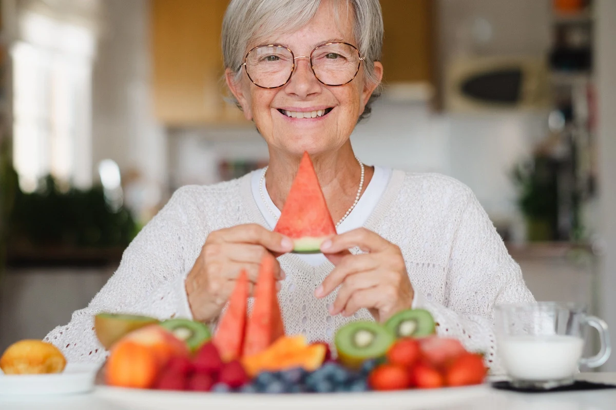 Senior Nutrition Tips – The Village Senior Living Smiling older adult holds a slice of watermelon at a table filled with fresh fruit, showing senior nutrition tips at The Village Senior Living.